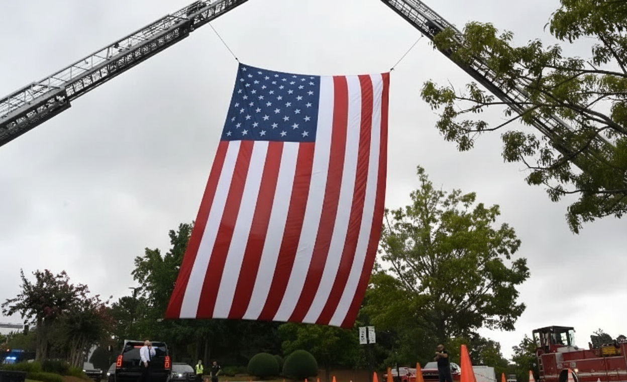 Large American flag suspended between two fire truck ladders during a public event.